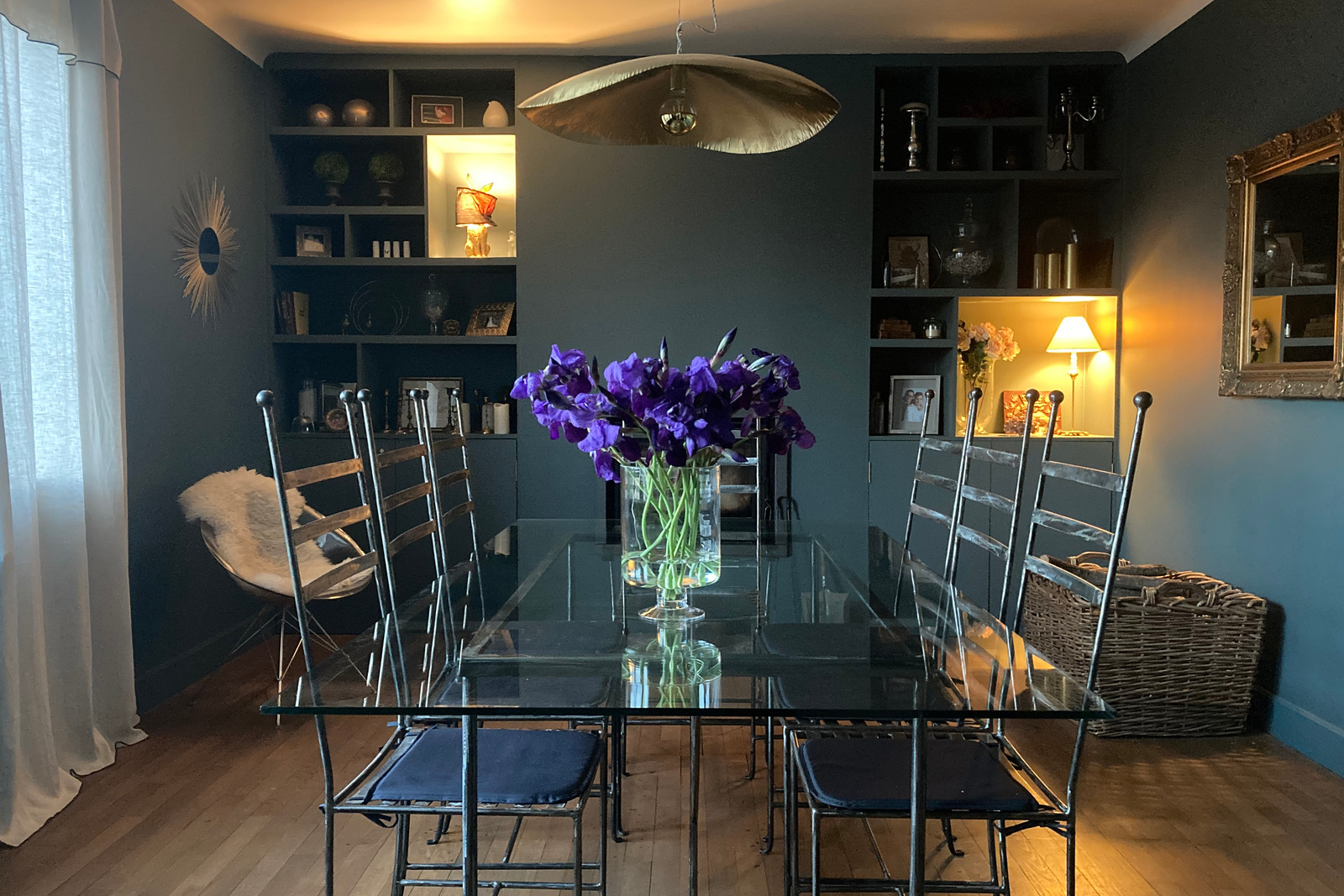 Table à manger en verre et chaise en métal à Blois, Armoire sur mesure couleur sombre en Loir et Cher, Lampe vintage allumée à Mesland, Miroir style vintage et moderne en or, Fleur violette en milieu de table 