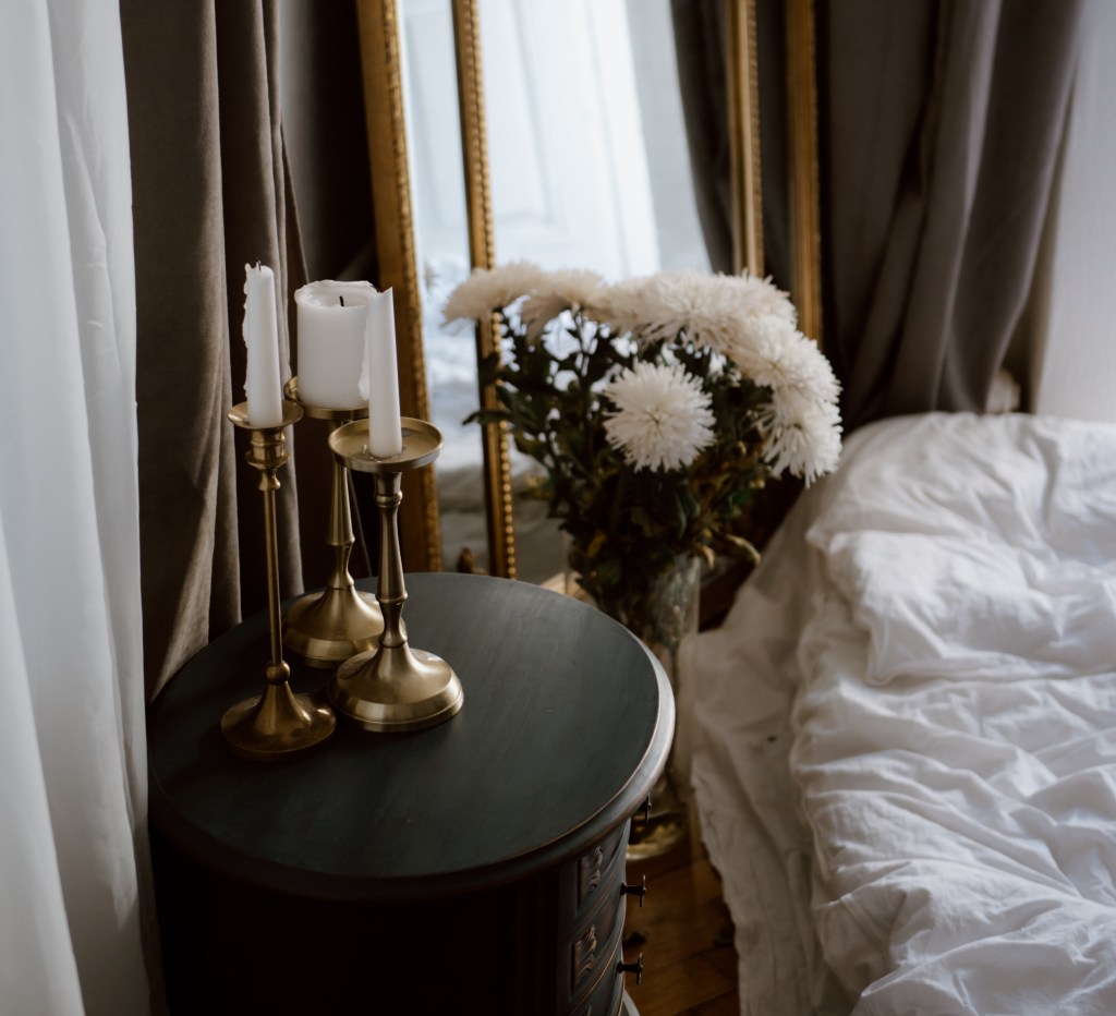 Table de chevet en bois noir à Blois, Fleur et bougie blanche en Loir et Cher, avec un miroir et des bougeoirs en or à Mesland,  Lit avec couette blanche rideau avec couleur foncée pour chambre,
